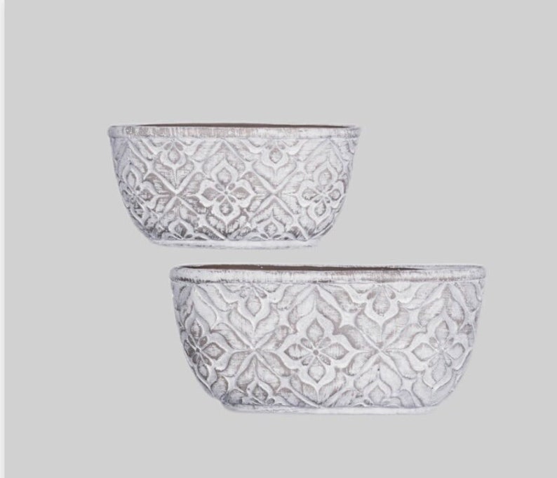 K&ouml;nnte beinhalten: Zwei wei&szlig;e ovale Pflanzgef&auml;&szlig;e mit einem gepr&auml;gten, filigranen Design. Die T&ouml;pfe sind aus einem strukturierten Material gefertigt und haben eine leicht abgenutzte Optik.