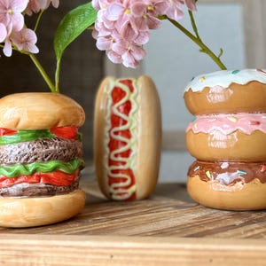 May include: Three ceramic food-themed vases shaped like a hamburger, a hot dog, and a stack of three donuts. The hamburger vase features lettuce, tomato, and meat layers. The hot dog vase has red and yellow drizzle. The donut vase has white, pink, and chocolate icing.