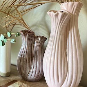 May include: Two ceramic vases with a vertical ribbed texture and ruffled rims. The larger vase is a pale pastel pink, while the smaller vase features a darker, brownish-pink glaze. Both vessels have a bulbous body shape and a glossy finish.
