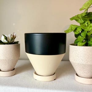 May include: Three ceramic planters of varying sizes. The smallest planter is filled with a white flower. The middle planter is black and cream. The largest planter holds a green plant. All planters have matching saucers.