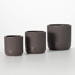 May include: Three dark gray ceramic planters of varying sizes. Each cylindrical pot features vertical ribbed detailing on the upper portion and a solid, darker base. The planters are designed for indoor or outdoor use.