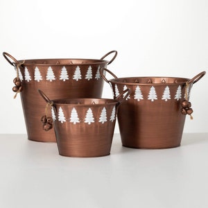 May include: Three copper-colored metal buckets with handles, varying in size. Each bucket features a white painted Christmas tree design around the rim. Small bells are attached to the handles with twine. Ideal for holiday decor.