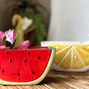 May include: Two ceramic fruit-shaped bowls. One is a watermelon slice, red with black seed details, holding pink flowers. The other is a lemon slice, yellow with white details. Both are on a wooden surface.