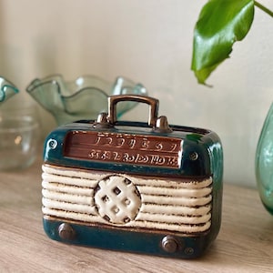 May include: A ceramic radio-shaped decorative object featuring a deep teal blue finish with a cream-colored ribbed speaker grille and a central circular pattern. The top includes a handle, and the front displays embossed numbers and radio dial markings.