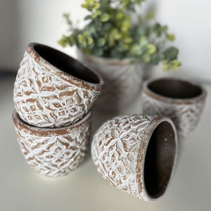 May include: Set of four white ceramic planters with intricate floral designs. The planters are stacked on top of each other, with the top planter slightly tilted to the side. The planters are made of a rough, textured material and have a rustic, farmhouse style.