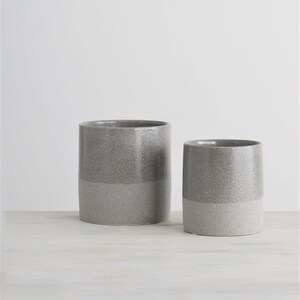 May include: Two gray ceramic planters with a speckled glaze. The larger planter is taller and wider than the smaller planter.