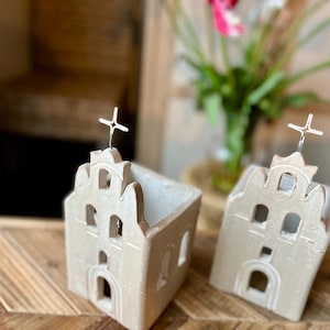 May include: Two beige ceramic church-shaped candle holders with crosses on top. The buildings have arched windows and doors. They are placed on a wooden surface, with a blurred background of flowers and a wicker chair.