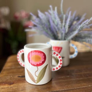 May include: Two white ceramic mugs with floral designs. One mug features a large pink and orange flower, while the other has a red flower. Both mugs have handles decorated with red flowers and blue polka dots.