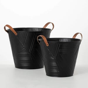 May include: Two black metal buckets with brown leather handles. The buckets have a textured geometric design. The handles are attached to the buckets with rivets. The buckets are of different sizes.