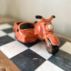 May include: A glossy orange ceramic motorcycle figurine with a sidecar. The motorcycle features a black front tire, black handlebars, and a white circular headlight. The sidecar has a black interior opening, and the entire piece is finished with a smooth, reflective glaze that highlights the curved, molded details of the vehicle's frame and body.