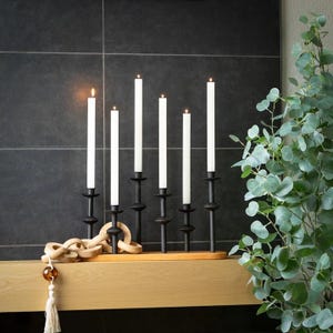May include: A black metal candelabra holds six white taper candles, some lit. The candelabra sits on a wooden shelf with a decorative wooden chain and a tassel. A leafy green plant is in the background.