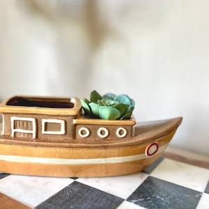May include: A ceramic boat-shaped planter in shades of tan and brown, with white accents and a red circle. The planter holds a green succulent. The boat has rectangular and circular details, and a white stripe along the hull.
