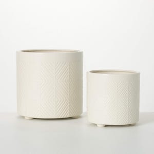May include: Two off-white ceramic planters. The larger planter is cylindrical with a geometric pattern and sits on three small feet. The smaller planter has a similar design. Both planters have a smooth, glossy finish and are empty.