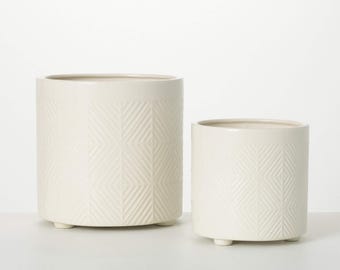 May include: Two off-white ceramic planters. The larger planter is cylindrical with a geometric pattern and sits on three small feet. The smaller planter has a similar design. Both planters have a smooth, glossy finish and are empty.