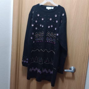 Può includere: Maglione nero con motivi geometrici decorativi e abbellimenti. Il maglione ha un girocollo e maniche lunghe. Il design include file di accenti bianchi, dorati e viola, creando un look unico. Il maglione è appeso a una gruccia bianca.