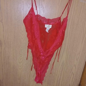 May include: A bright red lace bodysuit featuring thin adjustable shoulder straps, a deep V-neckline with scalloped lace trim, and a high-cut leg opening. The garment includes a white brand label at the neckline and delicate ribbon accents.