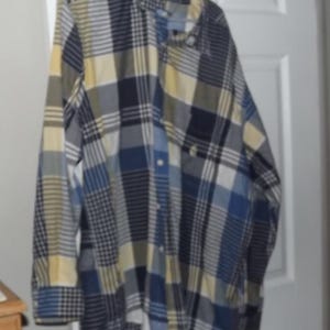 Puede incluir: Una camisa abotonada de manga larga con un estampado de cuadros en tonos azules, amarillos y grises. La camisa está colgada de una percha contra una puerta blanca. La camisa tiene cuello abotonado y bolsillo en el pecho.