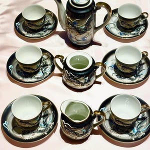 Op de afbeelding: Een vintage zwart en goud theeservies met drakenmotieven. De set bevat een theepot, een suikerpot, een melkkan en zes theekopjes met schotels. De kopjes en schotels hebben gouden randen. De theepot heeft een gouden handvat en een witte tuit.