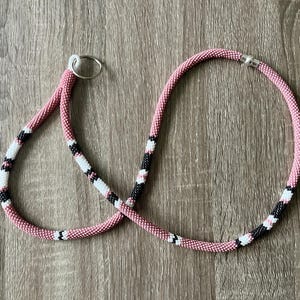 May include: A pink, black, and white beaded lanyard with a silver key ring. The lanyard is made of small beads and has a repeating pattern of pink, black, and white.