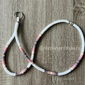 May include: White beaded lanyard with pink and purple accents, featuring a silver clasp and a silver ring at the top. The lanyard is on a woodgrain surface. @shoplanyardsbylaura