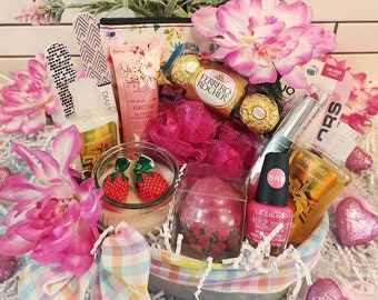 Può includere: Un cesto regalo pieno di articoli rosa e bianchi, tra cui una candela, una bomba da bagno, smalto per unghie, crema per le mani e cioccolatini. Il cesto è decorato con fiori rosa e cuori.