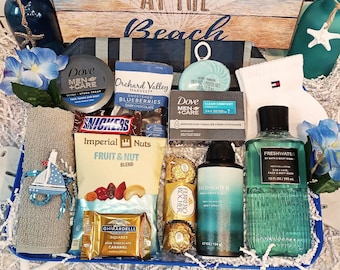 May include: A blue and white gift basket filled with various items, including a blue and green bottle of body wash, a bar of soap, a tube of body cream, a package of chocolate, a bag of nuts, a package of chocolate squares, a package of chocolate candies, a towel, and a pair of socks. The basket is decorated with a blue and white nautical theme.