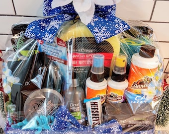 May include: A gift basket wrapped in clear cellophane, tied with a blue snowflake ribbon and a white flower. The basket contains car care products, Dove Men+Care products, and a Hershey's chocolate bar. Small gift boxes are at the base.