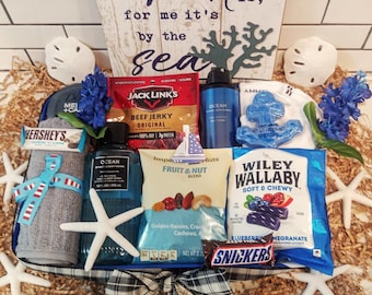 May include: A blue and white gift basket filled with various items, including a blue and white striped towel, a bottle of body wash, a bag of trail mix, a bag of gummy candies, a chocolate bar, and a sign that reads "heart is, for me it's by the sea".