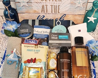 May include: A gift basket with a beach theme, containing a variety of items. Includes Dove Men+Care products, snacks like Snickers and nuts, and a bottle of Teakwood body wash. The basket is decorated with blue flowers and a wooden sign that says "Better at the Beach."