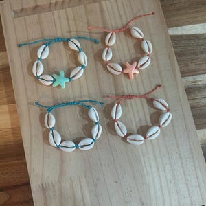May include: Four adjustable bracelets made with white cowrie shells strung on coloured cord. Two bracelets feature teal cord, one with a light blue starfish charm. Two bracelets feature coral cord, one with a light peach starfish charm.