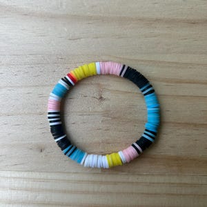 Puede incluir: Una pulsera colorida hecha de cuentas planas y redondas. Las cuentas son de varios colores, incluyendo negro, azul, rosa, amarillo, blanco y rojo. La pulsera es circular y parece estar hecha de un material elástico.