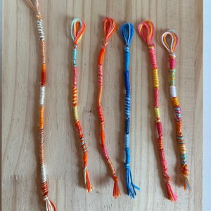 Op de afbeelding: Zes kleurrijke handgemaakte vriendschapsarmbanden worden getoond op een houten oppervlak. Elke armband heeft een lus aan de bovenkant en een kwast aan de onderkant, met verschillende tinten oranje, roze, geel, blauw en witte draad.