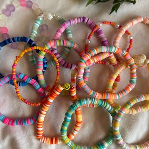 May include: A collection of colorful bracelets made with stacked, round beads. The bracelets feature a variety of colors, including pink, blue, green, yellow, and orange. Some bracelets have a smiley face charm.