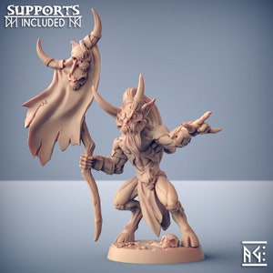 Op de afbeelding: Beige beeldje van een gehoornde demon met een versleten cape versierd met schedels. De demon houdt een staf vast en heeft een dreigende uitdrukking. De tekst "SUPPORTS INCLUDED" is bovenaan zichtbaar. Het beeldje staat op een beige voet.