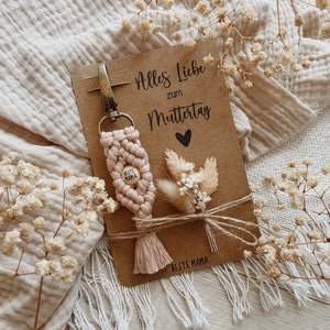Könnte beinhalten: Ein handgefertigter Makramee-Schlüsselanhänger in Hellrosa, mit einem Holzperlen-Schriftzug "BESTE MAMA". Dazu eine Karte mit dem Text "Alles Liebe zum Muttertag" und einem kleinen Trockenblumenarrangement, mit Bindfaden gebunden.