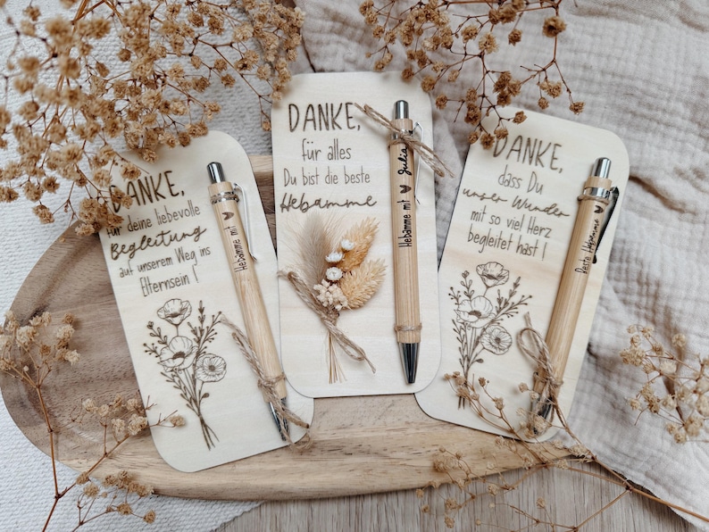 May include: Three wooden cards with engraved text and floral designs, each accompanied by a bamboo pen. The cards say "DANKE" in German, with additional text. Dried flowers and twine accents add a rustic touch to the presentation.