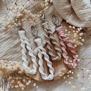 Könnte beinhalten: Vier Makramee-Schlüsselanhänger in Creme, Beige, Hellbeige und Rosa, jeweils mit einem silberfarbenen Verschluss. Die Schlüsselanhänger sind auf einem Holzbrett angeordnet, mit getrockneten weißen Blumen und einem beigen Stoff im Hintergrund. Die Schlüsselanhänger haben ein gewebtes Design.