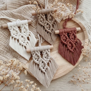 Könnte beinhalten: Vier Makramee-Wandbehänge in Creme-, Beige- und Terrakottafarben werden auf einem Holztablett präsentiert. Jedes Stück hat eine Holzleiste oben und ein rautenförmiges Design mit geknoteten Details und Fransen.