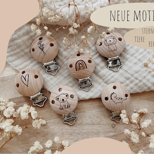 Könnte beinhalten: Holz-Schnullerclips mit verschiedenen Gravuren, darunter ein Herz, Blumen, ein Löwe, ein Regenbogen, ein Koala und ein Dinosaurier. Die Clips sind an Metallklammern befestigt. Das Bild enthält den Text "NEUE MOTIVE" und "STERNZEICHEN TIERE REGENBOGEN."