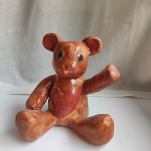 Peut inclure: Figurine d'ours en peluche en céramique faite à la main. L'ours est brun avec un détail en forme de cœur rouge sur sa poitrine. Il a de petits yeux noirs et est posé avec un bras levé. La figurine est assise sur une surface blanche.