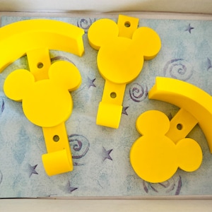 Pode incluir: Três ganchos de parede de plástico amarelo em forma de cabeça do Mickey Mouse. Os ganchos estão em uma caixa com um fundo estampado azul e branco.