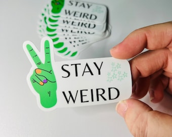 May include: A white sticker with a green peace sign and the text "STAY WEIRD" in black. The peace sign has colorful nail polish on the fingers. There are small green stars in the top right corner of the sticker.