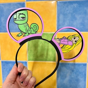 Peut inclure: Une paire d'oreilles de souris roses avec un bandeau noir. Une oreille représente un caméléon vert avec une expression triste. L'autre oreille représente une princesse blonde avec une robe rose et de longs cheveux.
