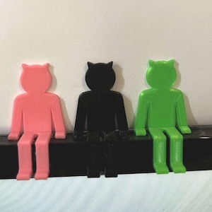 Può includere: Tre figurine di persone stampate in 3D sedute su una superficie nera. Le figurine sono rosa, nera e verde.