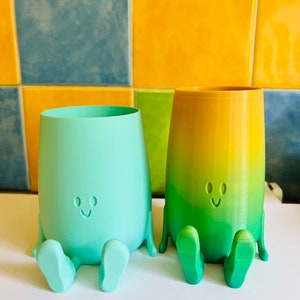 May include: Two whimsical, smiling planters in the image. One is a solid mint green, the other is a gradient of yellow and green. Both have legs and feet, and are sitting. The background is a colorful tile wall.