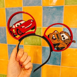 Könnte beinhalten: Rotes und schwarzes Mickey Mouse Ohrenband mit einem roten Auto und einem braunen LKW auf den Ohren.