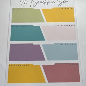 Könnte beinhalten: Ein farbenfroher Wochenplaner mit dem Text "Ant Seachtain Seo" oben. Jeder Wochentag wird durch eine andere Farbe dargestellt, darunter Grün, Gelb, Türkis, Rosa, Lila, Blau und Gold. In der unteren rechten Ecke steht das Wort "NOTAI".