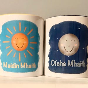 以下が含まれることがあります： カラフルなイラストが描かれた2つの白いセラミックマグ。1つのマグには「Maidin Mhaith」の文字と笑顔の太陽、黄色の持ち手があります。もう1つのマグには「Oíche Mhaith」の文字と笑顔の月、黒い持ち手があります。