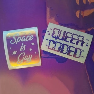 May include: Two iridescent stickers with white text. One sticker says "Space is Gay" with a stylized font and stars. The other sticker says "QUEER CODED" in a pixelated font.