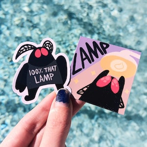 Könnte beinhalten: Zwei schwarze und rote Motten-Aufkleber. Ein Aufkleber hat den Text "100% THAT LAMP" darauf. Der andere Aufkleber hat den Text "LAMP" in großen Buchstaben.